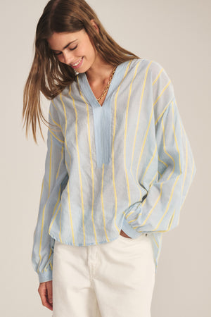 A woman wears the ANALYN TOP by Velvet by Graham & Spencer—a relaxed fit, V-neck, yellow yarn-dyed stripe cotton top—paired with white pants, standing against a neutral background, looking down and smiling.