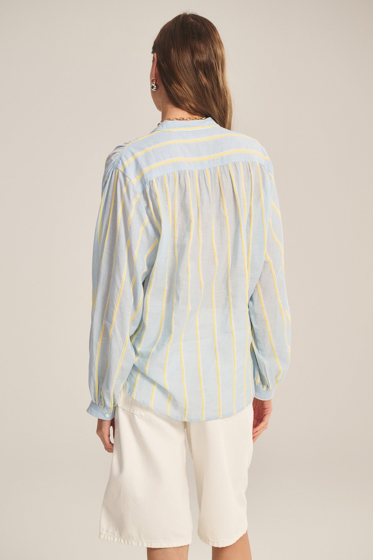 Person standing with their back to the camera, wearing the ANALYN TOP by Velvet by Graham & Spencer—a relaxed fit, light blue top with yellow yarn-dyed stripes—and white knee-length shorts.-40959849005249