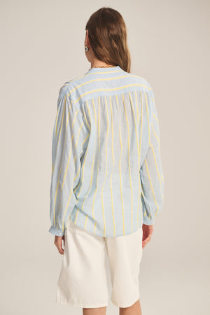 Person standing with their back to the camera, wearing the ANALYN TOP by Velvet by Graham & Spencer—a relaxed fit, light blue top with yellow yarn-dyed stripes—and white knee-length shorts.