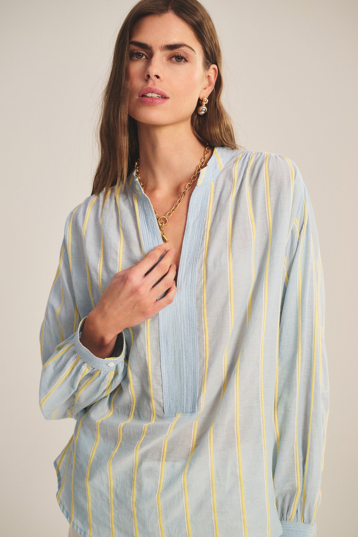 A woman wears the ANALYN TOP by Velvet by Graham & Spencer—a light blue, V-neck blouse with yellow vertical stripes, crafted from yarn-dyed stripe cotton—styled with gold jewelry against a neutral background.-40959849038017