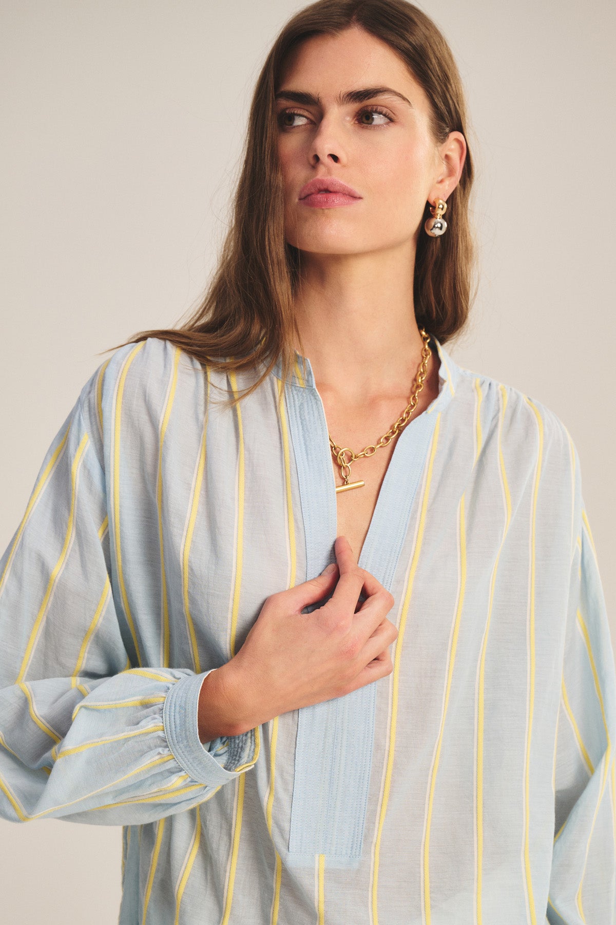 A woman wears the ANALYN TOP by Velvet by Graham & Spencer, a relaxed-fit V-neck in light blue and yellow yarn-dyed striped cotton, with a gold necklace and matching earrings, standing against a neutral background.-40959849070785