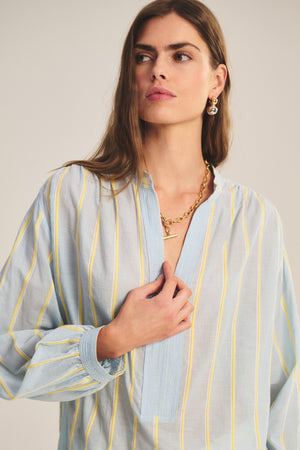 A woman wears the ANALYN TOP by Velvet by Graham & Spencer, a relaxed-fit V-neck in light blue and yellow yarn-dyed striped cotton, with a gold necklace and matching earrings, standing against a neutral background.