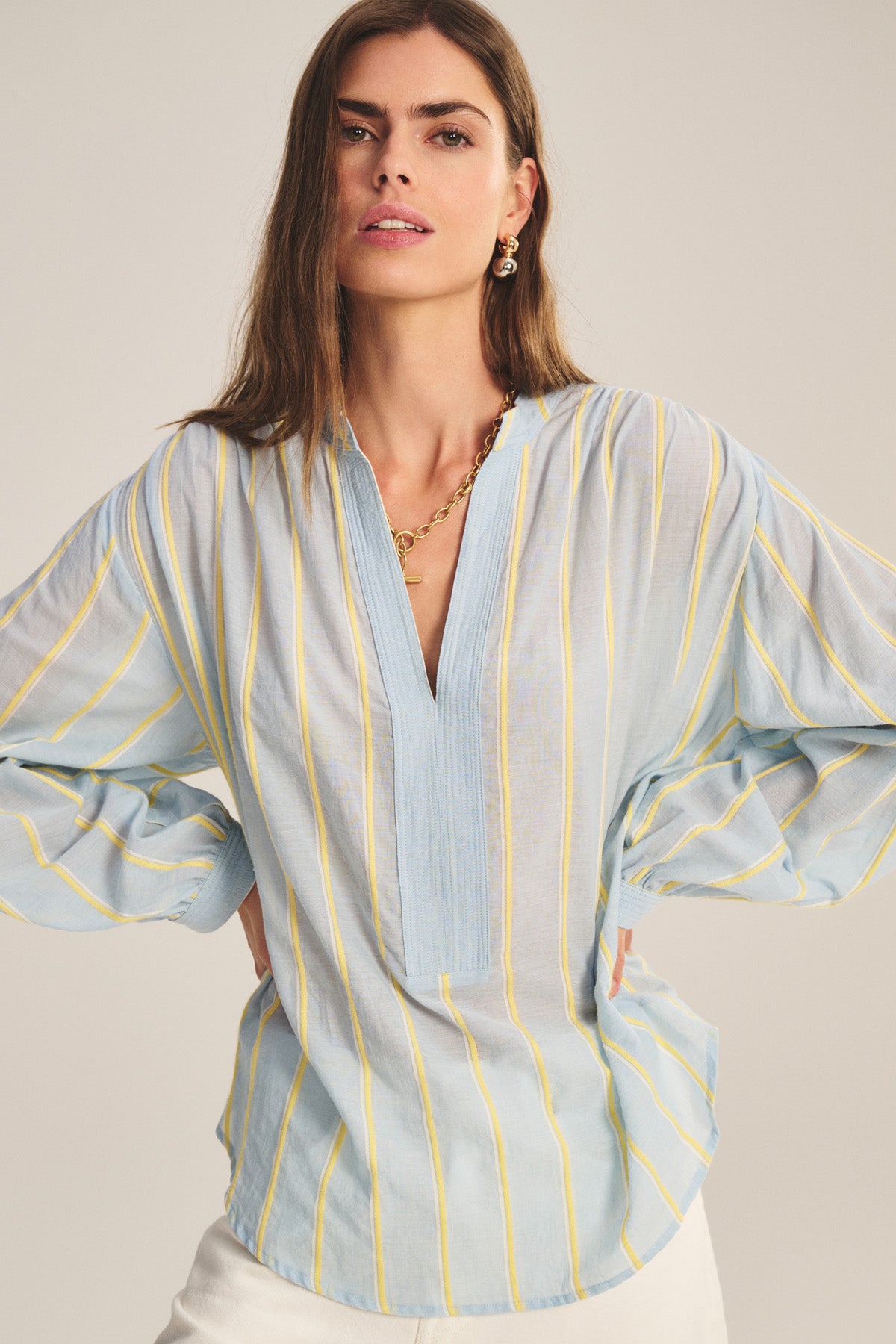 A woman stands with hands on hips against a neutral background, wearing the Velvet by Graham & Spencer ANALYN TOP—a light blue, yarn-dyed stripe cotton relaxed fit top with yellow vertical stripes and a V-neckline—paired with white pants.-40959849103553