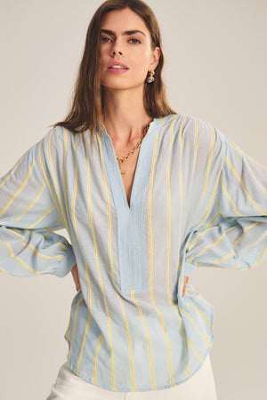 A woman stands with hands on hips against a neutral background, wearing the Velvet by Graham & Spencer ANALYN TOP—a light blue, yarn-dyed stripe cotton relaxed fit top with yellow vertical stripes and a V-neckline—paired with white pants.