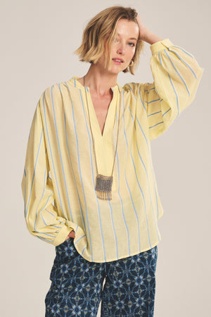 Wearing the Velvet by Graham & Spencer ANALYN TOP, a relaxed fit, yarn-dyed stripe cotton shirt, paired with patterned blue pants and a pendant necklace, this person stands with one hand on their head.