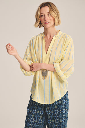 A woman wears the Velvet by Graham & Spencer ANALYN TOP—relaxed fit, yarn-dyed yellow and blue striped cotton—paired with blue patterned pants, standing against a neutral background.