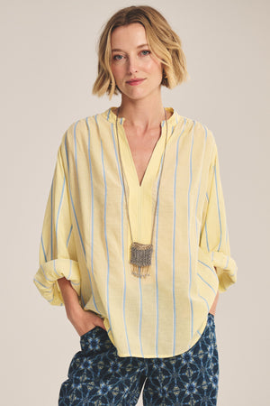 Wearing the ANALYN TOP by Velvet by Graham & Spencer—a relaxed fit V-neck in yellow and white stripe cotton with rolled-up sleeves—this person pairs it with patterned blue pants, a long pendant necklace, and stands hands in pockets.