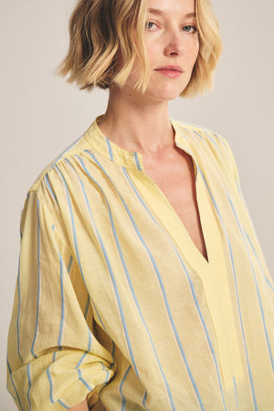 A person with short blonde hair wears the Velvet by Graham & Spencer ANALYN TOP—a relaxed fit, V-neck style in blue and white yarn-dyed stripe cotton with a hint of yellow.