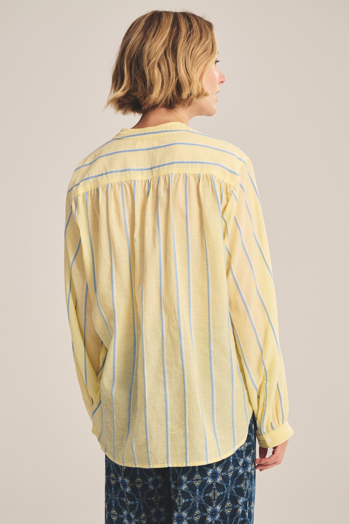 A woman with short blonde hair is seen from the back wearing the ANALYN TOP by Velvet by Graham & Spencer—a relaxed fit, loose yellow button-up shirt with blue and white yarn-dyed stripes—paired with patterned blue pants.
