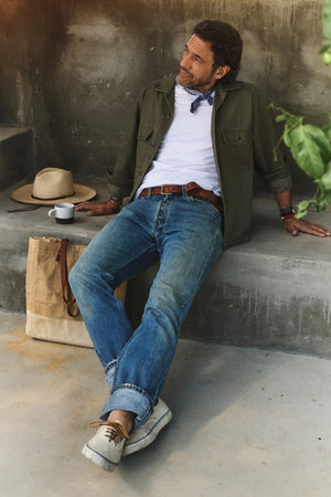 A man in jeans, a white tee, and the ARCHER SHIRT by Velvet by Graham & Spencer sits on a concrete bench beside a hat, bag, and coffee cup, gazing thoughtfully to the side.