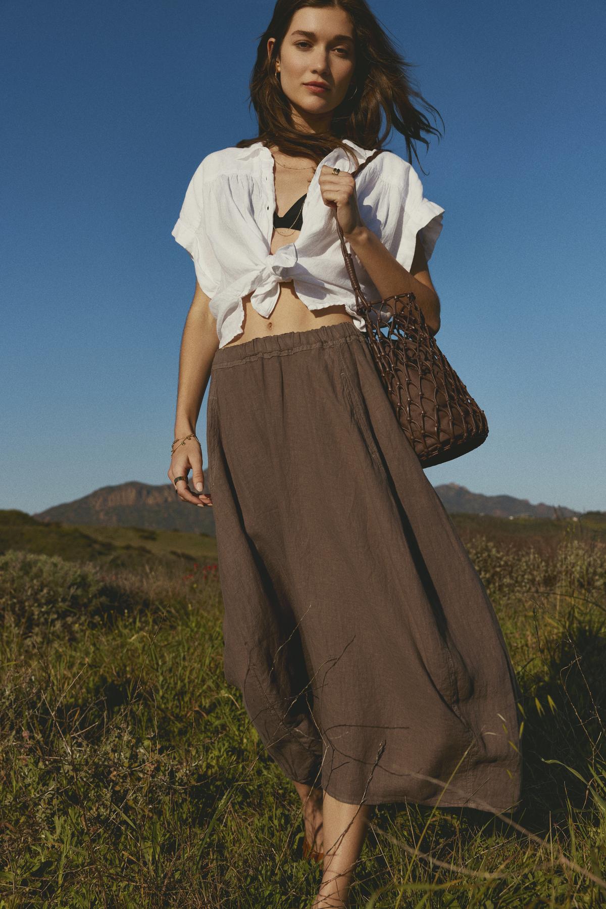   Woman in a white tied-up shirt and FAE LINEN A-LINE SKIRT with an elastic waistband standing in a field by Velvet by Graham & Spencer. 