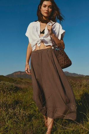 Woman in a white tied-up shirt and FAE LINEN A-LINE SKIRT with an elastic waistband standing in a field by Velvet by Graham & Spencer.