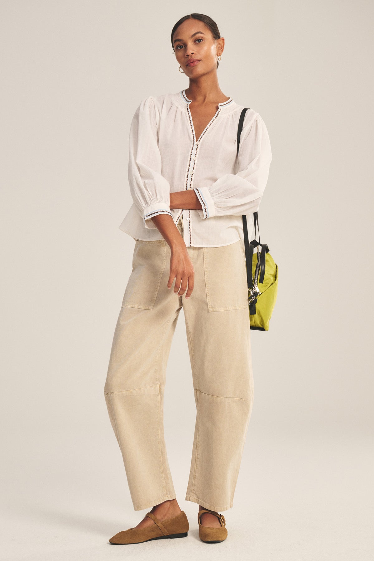 A woman wears the ARIANA TOP by Velvet by Graham & Spencer, a white long-sleeve blouse with smock detailing, styled with beige wide-leg pants, brown shoes, and a small yellow shoulder bag.