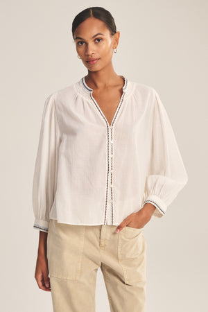 A person wears the ARIANA TOP by Velvet by Graham & Spencer—a white, long-sleeve blouse in embroidered cotton voile—with beige pants, standing against a neutral background.
