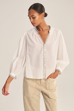 Model wears the ARIANA TOP by Velvet by Graham & Spencer—a loose, white, long-sleeve blouse with smock detailing and subtle embroidered trim—paired with light beige pants against a plain background.