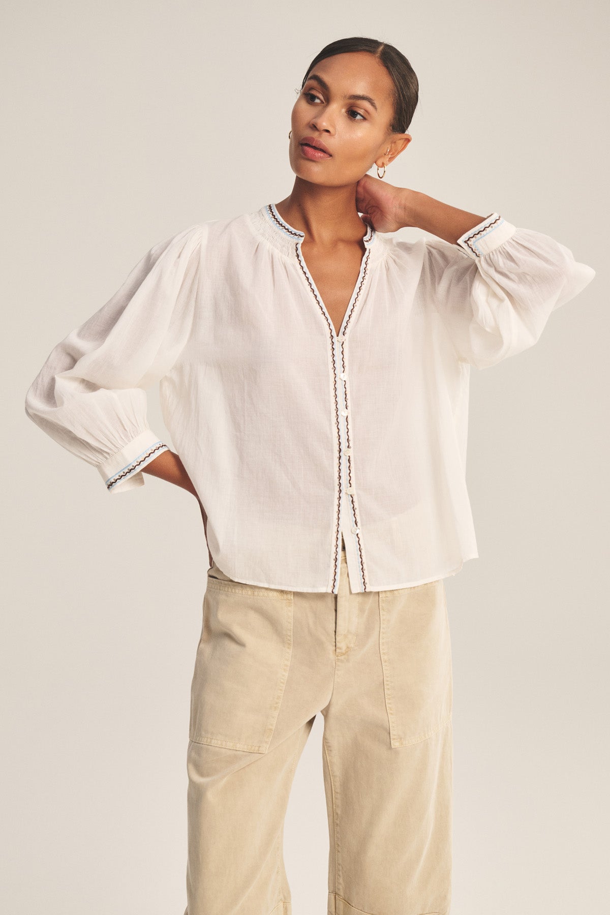A woman models the ARIANA TOP by Velvet by Graham & Spencer, a loose white cotton voile blouse with smock details and embroidered trim, styled with beige pants. She poses with one hand on her neck and the other in her pocket.-41080131092673
