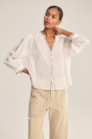A woman models the ARIANA TOP by Velvet by Graham & Spencer, a loose white cotton voile blouse with smock details and embroidered trim, styled with beige pants. She poses with one hand on her neck and the other in her pocket.
