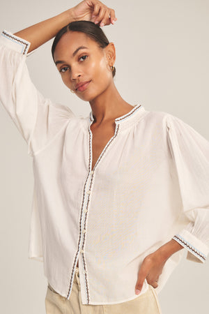 A woman poses with one arm raised, wearing the ARIANA TOP by Velvet by Graham & Spencer—a loose, white, long-sleeve blouse crafted from embroidered cotton voile with delicate trim—against a plain background.