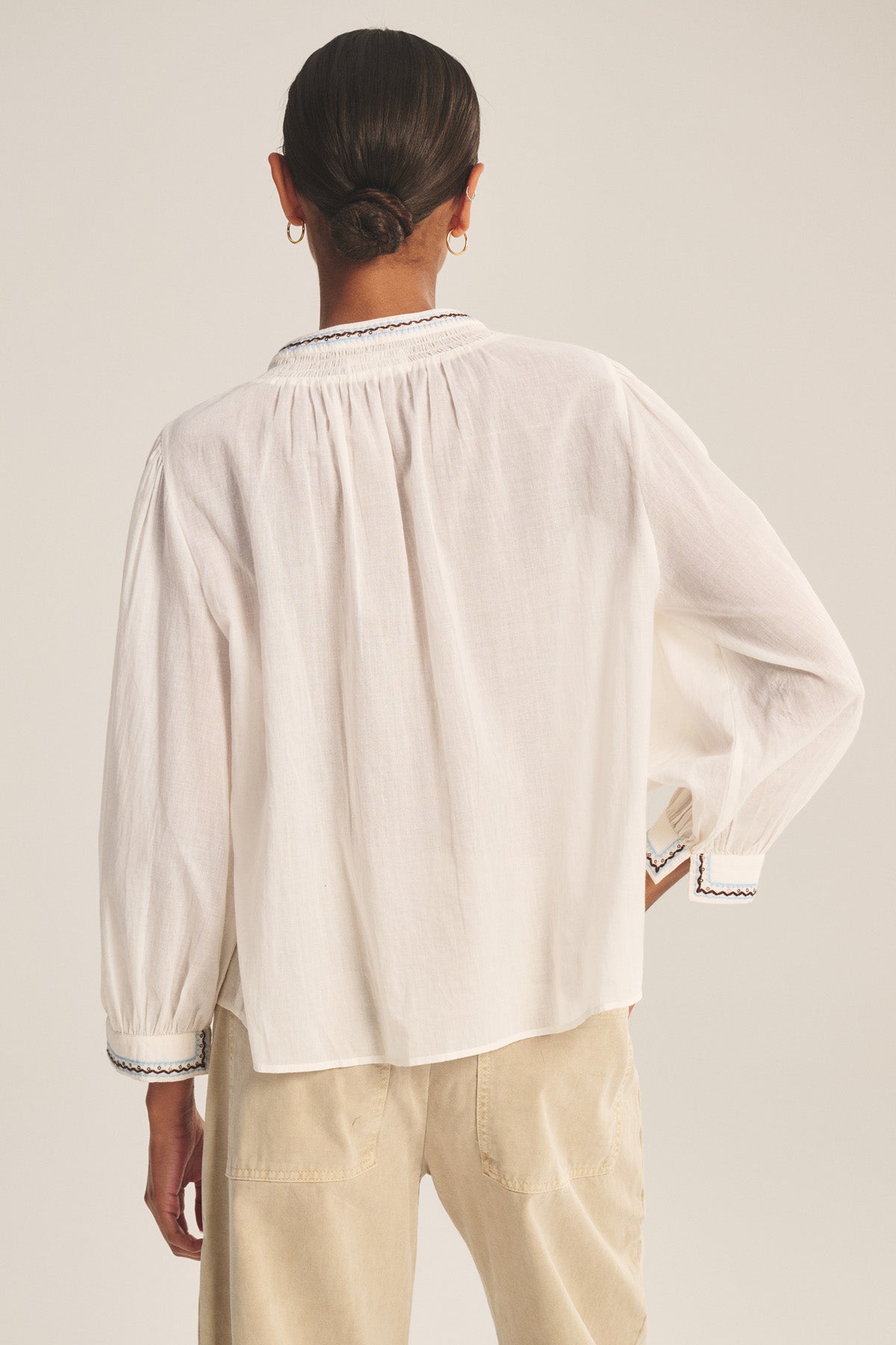Person with hair in a low bun wears the ARIANA TOP by Velvet by Graham & Spencer—a loose-fitting white long-sleeve blouse with smock detailing—paired with beige pants, shown from the back against a plain background.