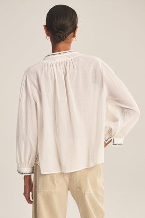Person with hair in a low bun wears the ARIANA TOP by Velvet by Graham & Spencer—a loose-fitting white long-sleeve blouse with smock detailing—paired with beige pants, shown from the back against a plain background.