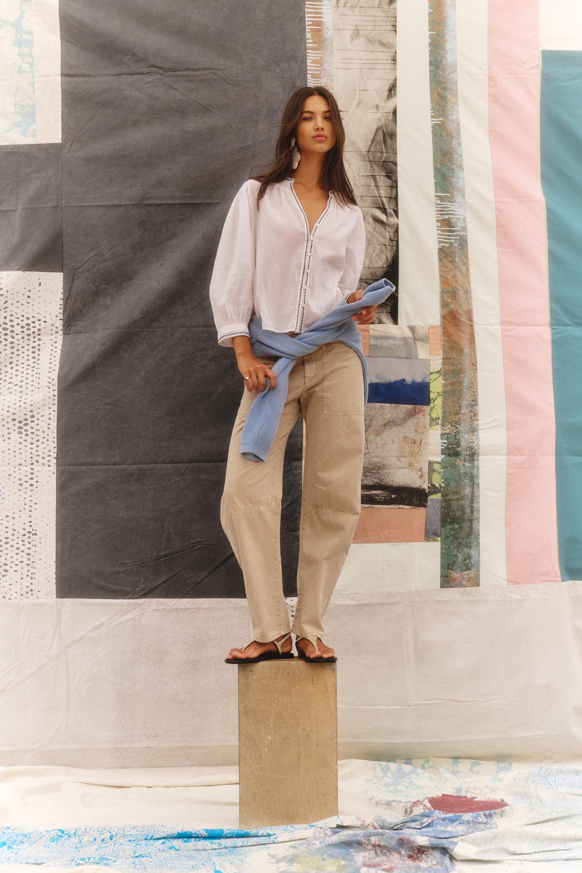 A woman stands on a wooden block wearing the ARIANA TOP by Velvet by Graham & Spencer with beige pants, sandals, and a light blue sweater around her waist, set against a backdrop of abstract art panels.-41080131223745