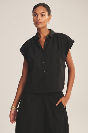 A woman wears the ARLEND TOP by Velvet by Graham & Spencer—a black cotton poplin cap-sleeve blouse—paired with a matching skirt, standing with one hand in her pocket against a neutral background.
