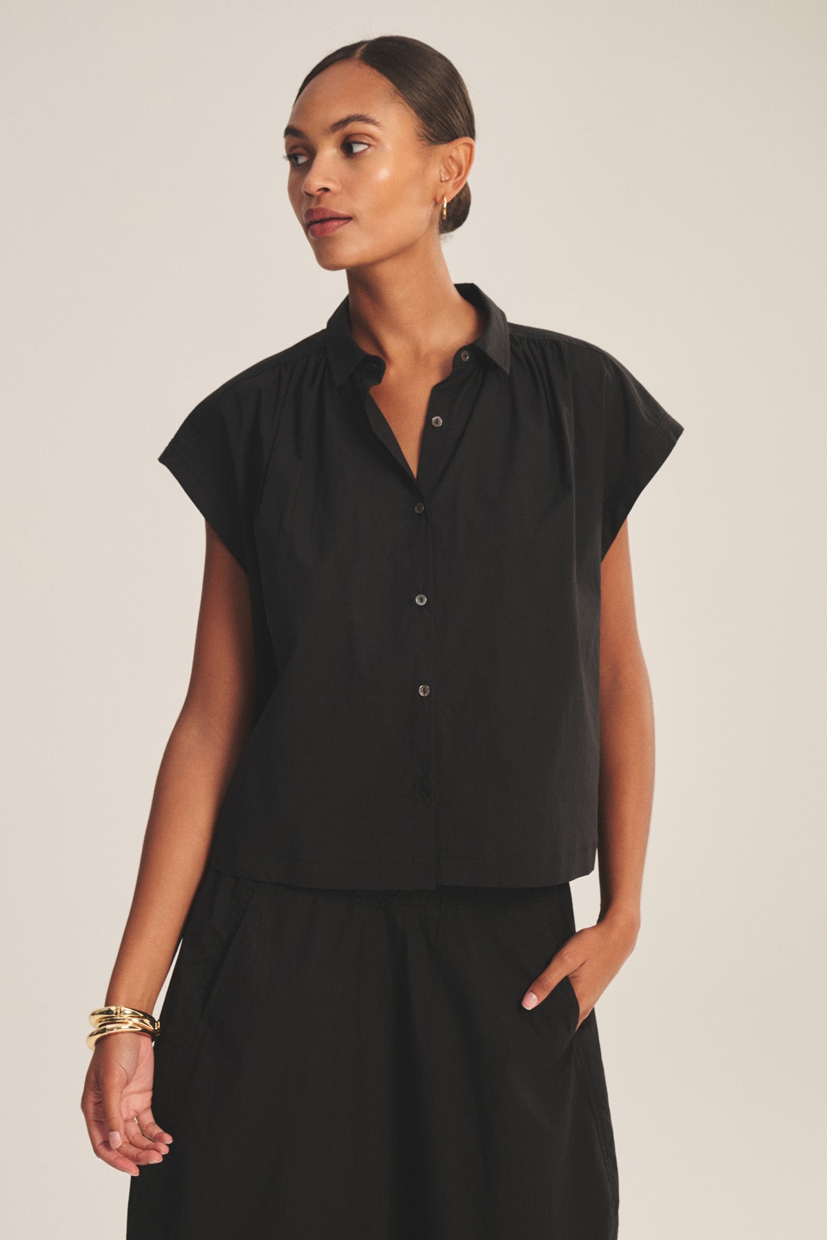A woman wears the ARLEND TOP by Velvet by Graham & Spencer—a black cotton poplin cap-sleeve top—paired with a matching black skirt, standing with one hand in her pocket and looking to the side against a plain background.-41037939343553