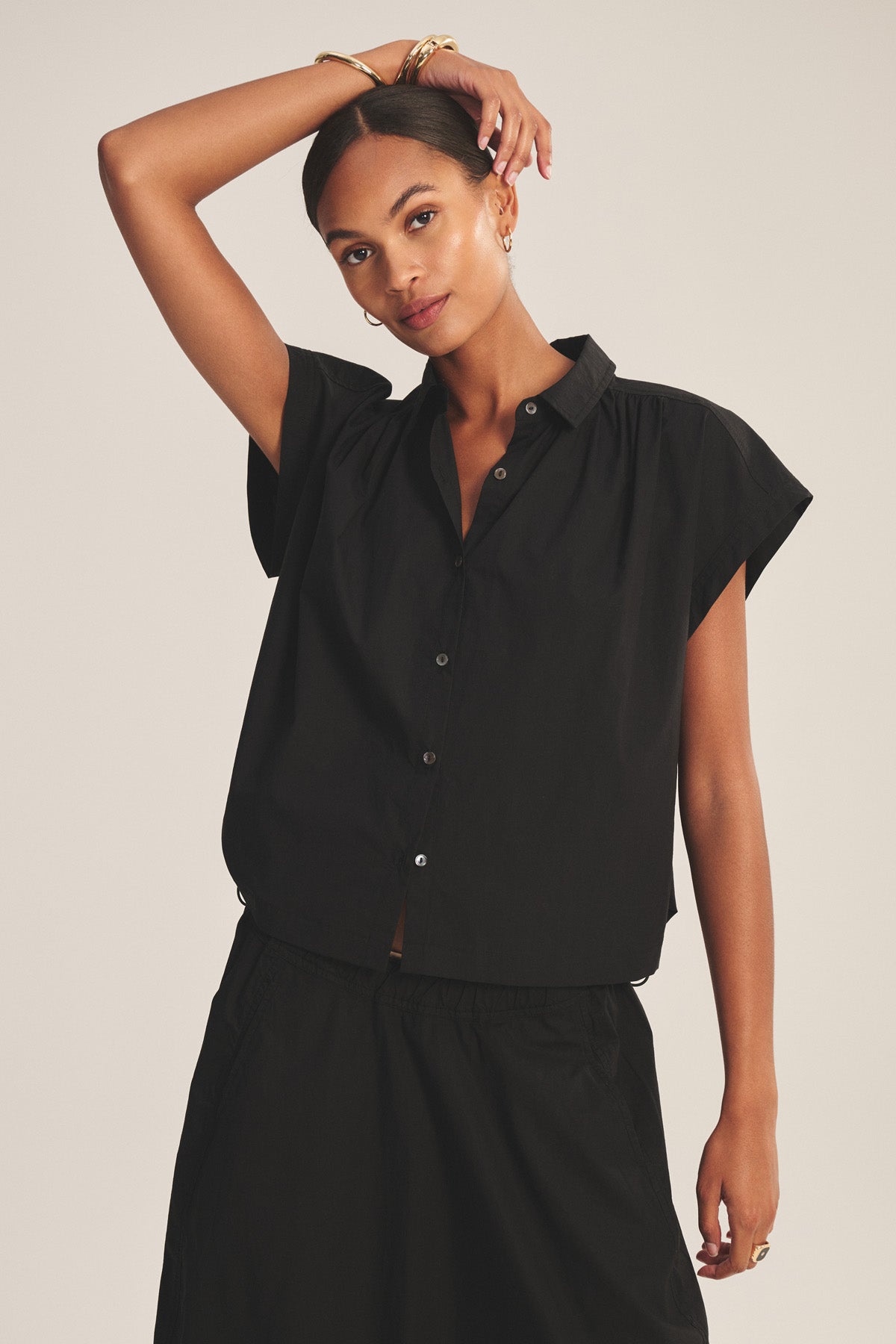 A woman poses with one arm raised and the other relaxed by her side, wearing the ARLEND TOP by Velvet by Graham & Spencer—a short-sleeved cotton poplin top—paired with a matching black skirt.-41037939409089