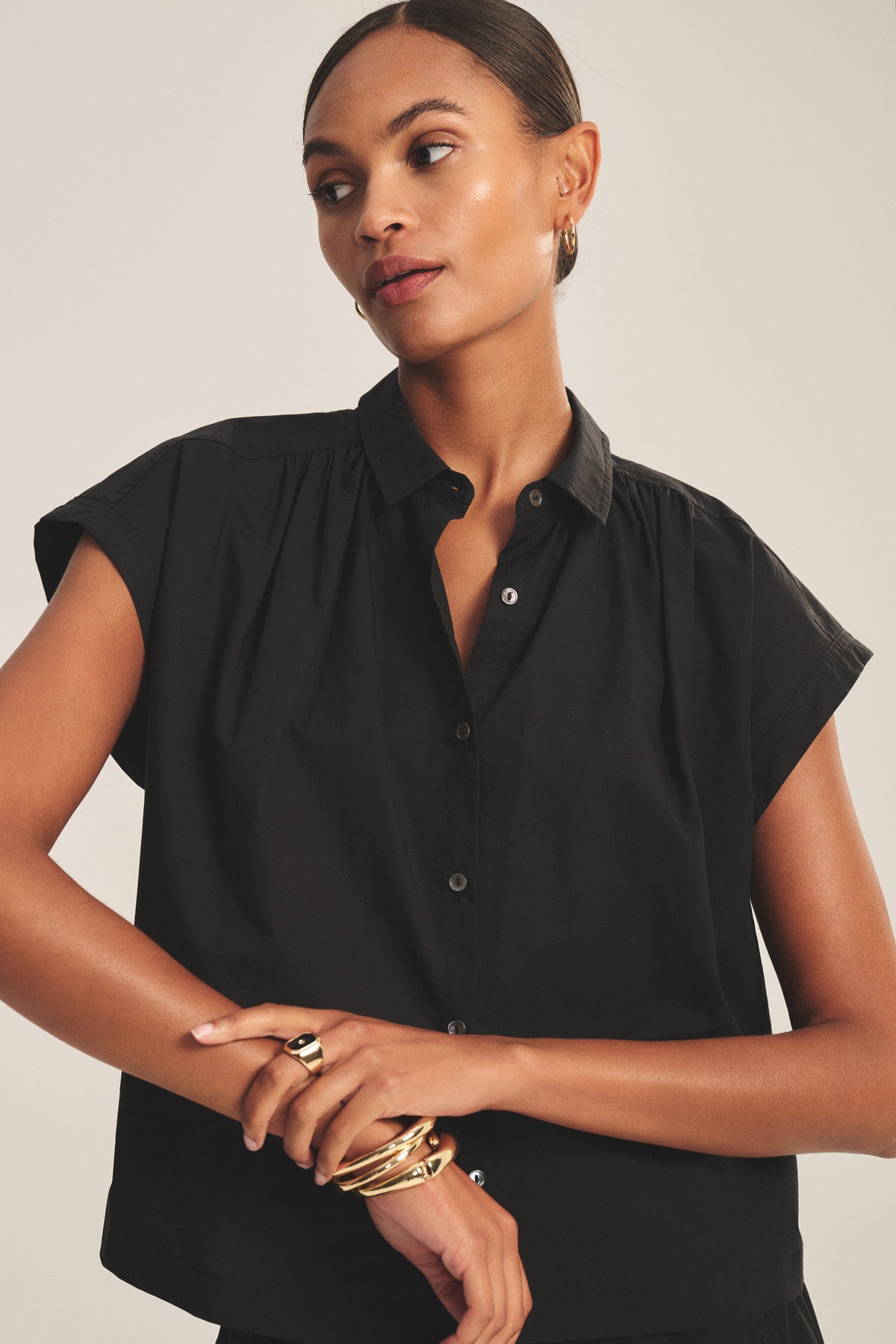 A woman with slicked-back hair models the ARLEND TOP by Velvet by Graham & Spencer—a black cotton poplin cap-sleeve top—paired with gold jewelry, standing against a plain light background.-41037939441857