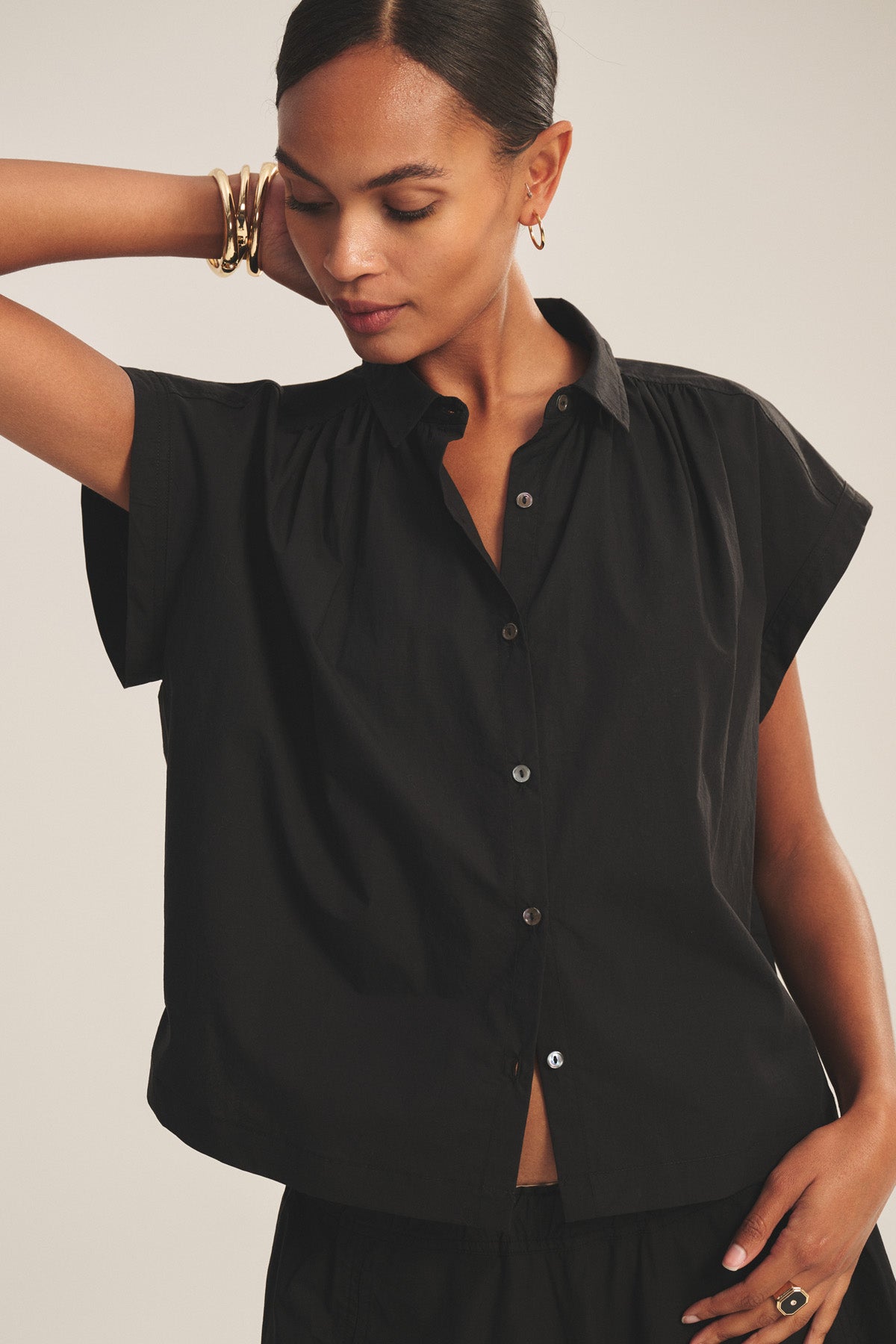 A woman wearing the ARLEND TOP by Velvet by Graham & Spencer—a short-sleeve black cotton poplin blouse—pairs it with gold jewelry, gazing downward with one arm raised behind her head against a simple backdrop.