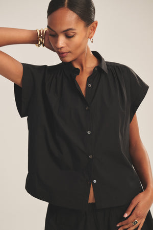 A woman wearing the ARLEND TOP by Velvet by Graham & Spencer—a short-sleeve black cotton poplin blouse—pairs it with gold jewelry, gazing downward with one arm raised behind her head against a simple backdrop.