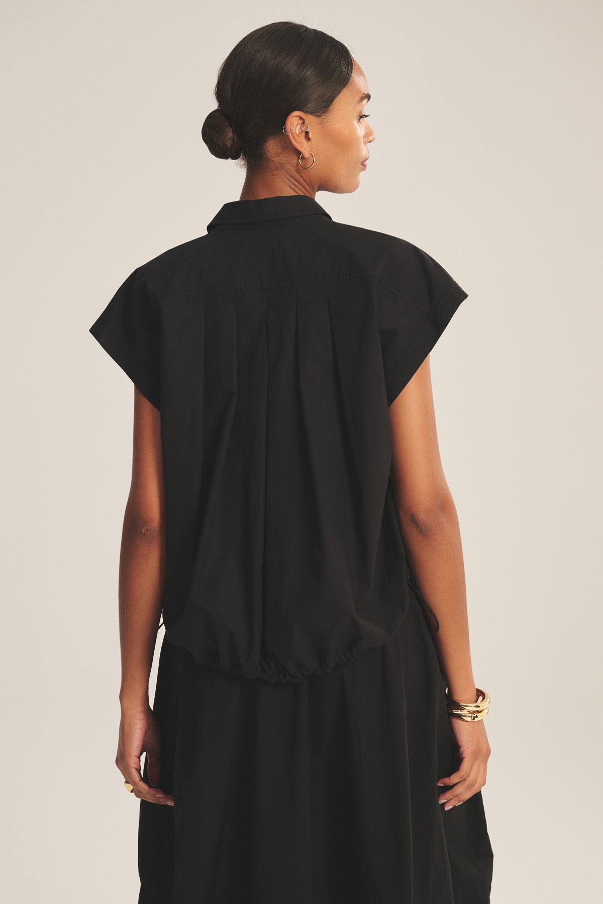 A woman faces away, wearing the ARLEND TOP by Velvet by Graham & Spencer in black cotton poplin with a skirt. Her hair is tied back and she accessorizes with gold hoop earrings and bracelets.-41037939540161
