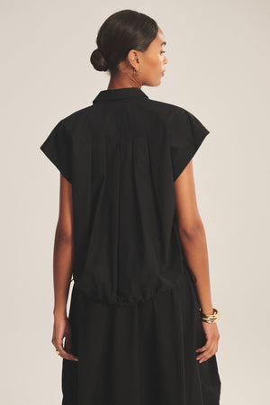 A woman faces away, wearing the ARLEND TOP by Velvet by Graham & Spencer in black cotton poplin with a skirt. Her hair is tied back and she accessorizes with gold hoop earrings and bracelets.
