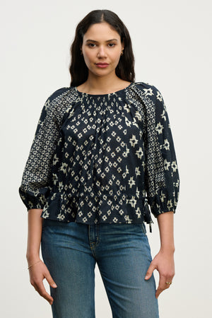 A woman stands against a plain background wearing the ADELAIDE TOP by Velvet by Graham & Spencer, a smocked cotton blouse with a white geometric pattern, ¾ sleeves, paired with blue jeans.