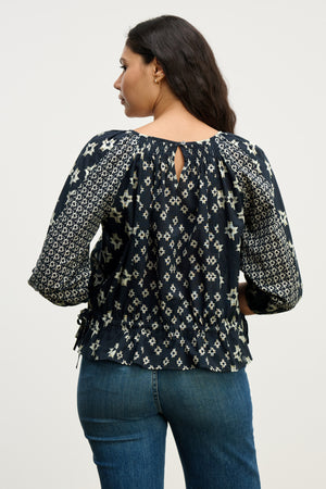 A woman with long dark hair is seen from behind, wearing the Velvet by Graham & Spencer ADELAIDE TOP in navy blue with ¾ sleeves and blue jeans against a plain background.