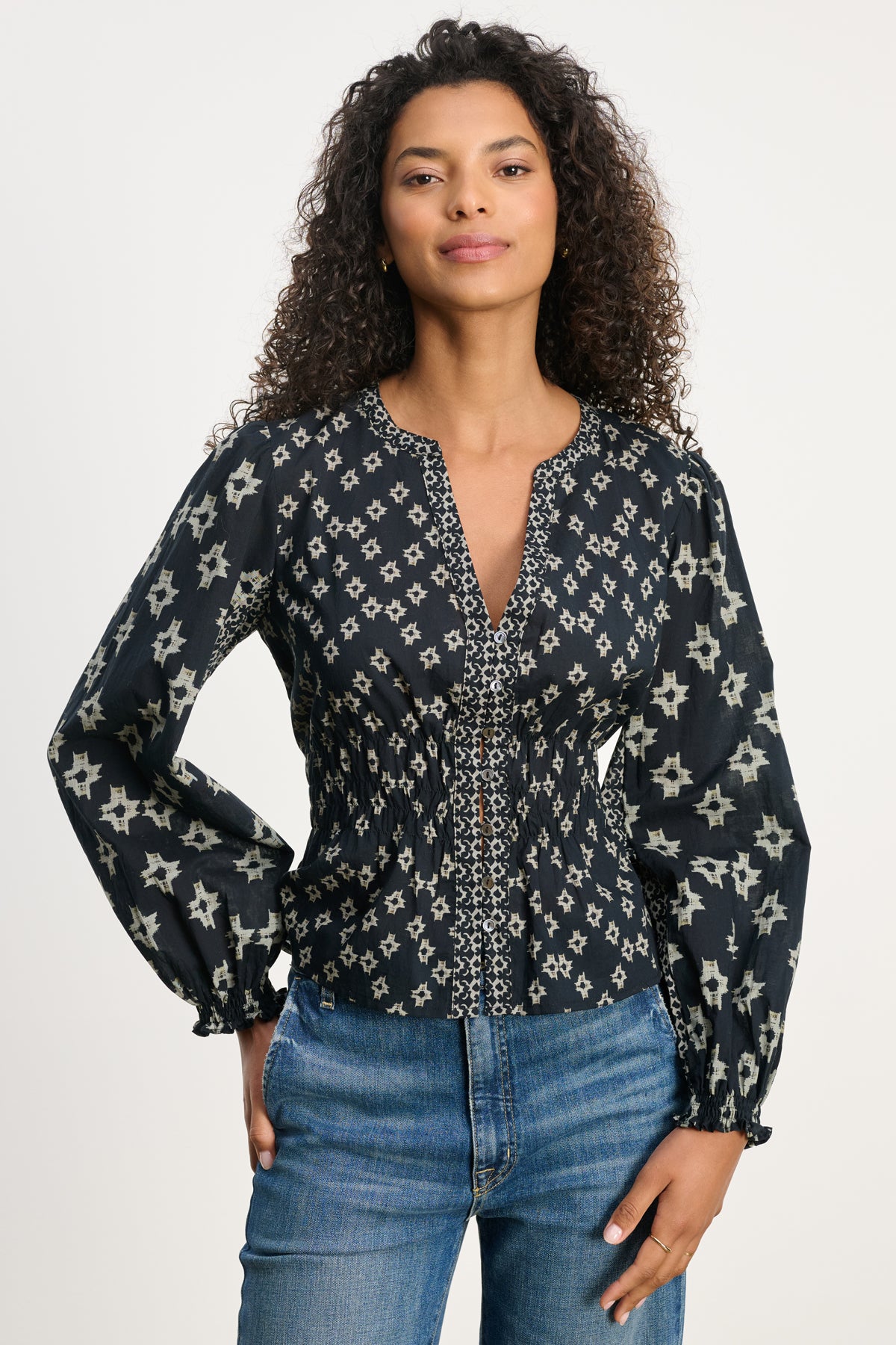   A person with curly hair wears the CHARLOTTE TOP by Velvet by Graham & Spencer—a long-sleeve, patterned black blouse with smocked elastic cuffs—and blue jeans, standing against a plain light background. 