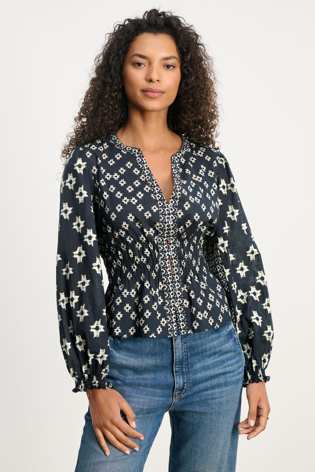 A woman with curly hair models the CHARLOTTE TOP by Velvet by Graham & Spencer—a dark blouse with smocked elastic cuffs and white star designs—paired with blue jeans against a plain light background.-39815174258881