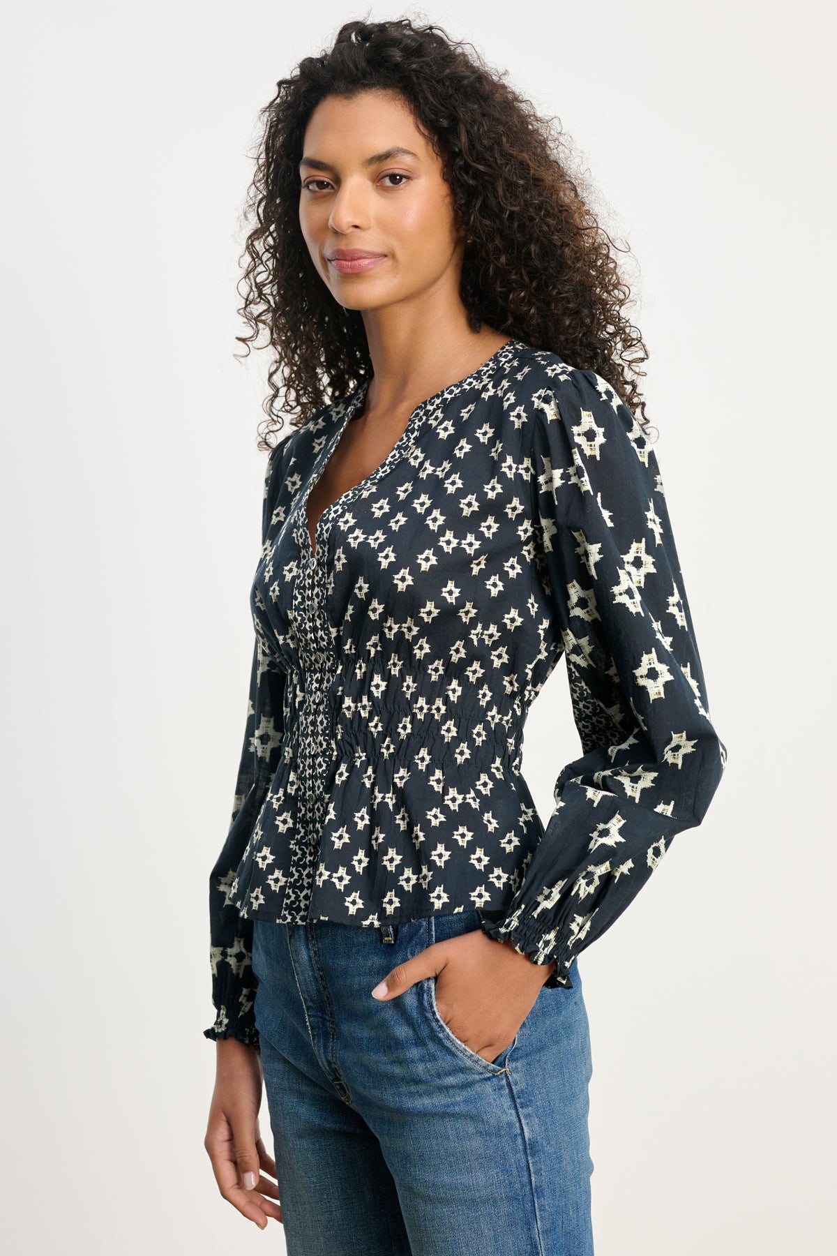A woman with curly hair models the Velvet by Graham & Spencer CHARLOTTE TOP—a dark patterned V-neck blouse—paired with blue jeans, standing with one hand in her pocket against a plain background.-39815174226113