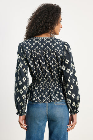 A woman with curly hair is shown from the back, wearing the Velvet by Graham & Spencer CHARLOTTE TOP—a dark long-sleeve blouse with smocked cuffs and a white star pattern—paired with blue jeans against a plain background.