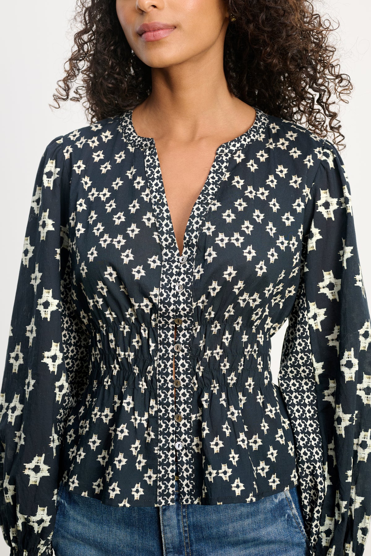  A woman wears the CHARLOTTE TOP by Velvet by Graham & Spencer—a navy blouse with white star prints, breathable cotton, smocked cuffs, V-neckline, button-down front, cinched waist, and long sleeves—paired with blue jeans. 