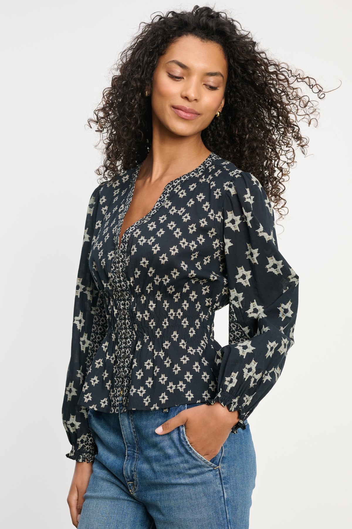 A woman with curly hair wears the Velvet by Graham & Spencer CHARLOTTE TOP—a dark, long-sleeve blouse with a light star pattern, smocked cuffs, and V-neckline—paired with blue jeans against a plain background.-39815174095041