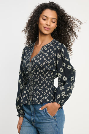 A woman with curly hair wears the Velvet by Graham & Spencer CHARLOTTE TOP—a dark, long-sleeve blouse with a light star pattern, smocked cuffs, and V-neckline—paired with blue jeans against a plain background.