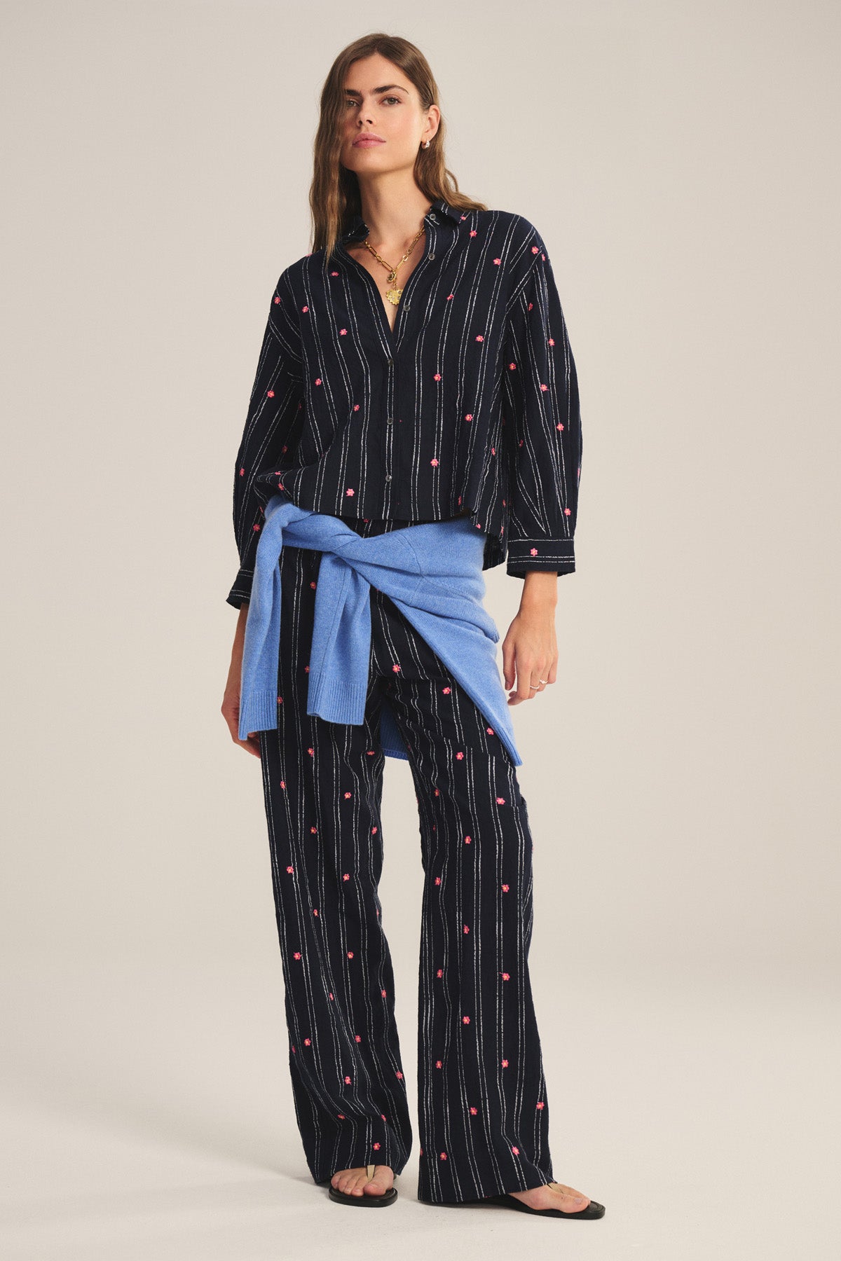 A woman wears the ASTREN SHIRT by Velvet by Graham & Spencer, a black pinstripe embroidered top with small red details, paired with matching pants, a blue sweater tied at the waist, and black sandals.-41284189126849