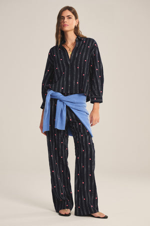 A woman wears the ASTREN SHIRT by Velvet by Graham & Spencer, a black pinstripe embroidered top with small red details, paired with matching pants, a blue sweater tied at the waist, and black sandals.