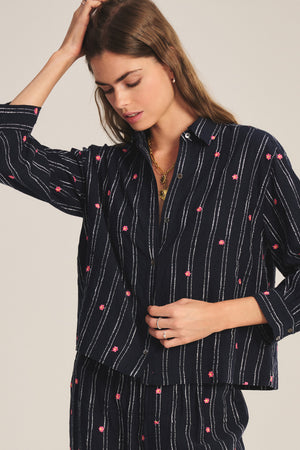 A woman wearing the ASTREN SHIRT by Velvet by Graham & Spencer: a navy blue, long-sleeve, button-up shirt with white stripes and pink floral prints, paired with matching pants.