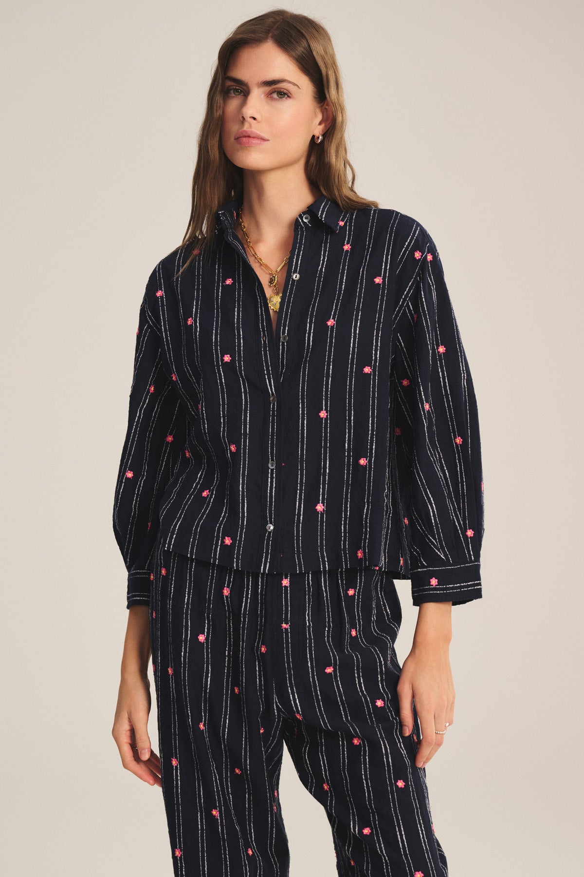 A woman wears the ASTREN SHIRT by Velvet by Graham & Spencer—a dark blue embroidered top with white stripes and pink dot patterns—paired with matching pants, standing against a plain beige background.-41284188963009