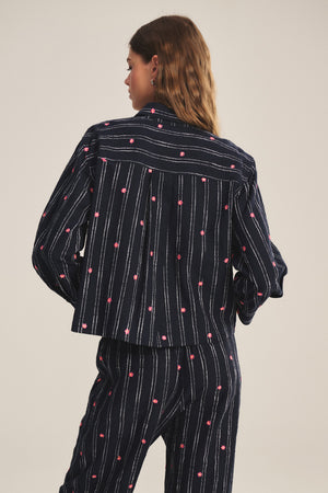 A woman stands with her back to the camera, wearing the ASTREN SHIRT by Velvet by Graham & Spencer—a cropped silhouette with vertical white stripes and small pink dot patterns.