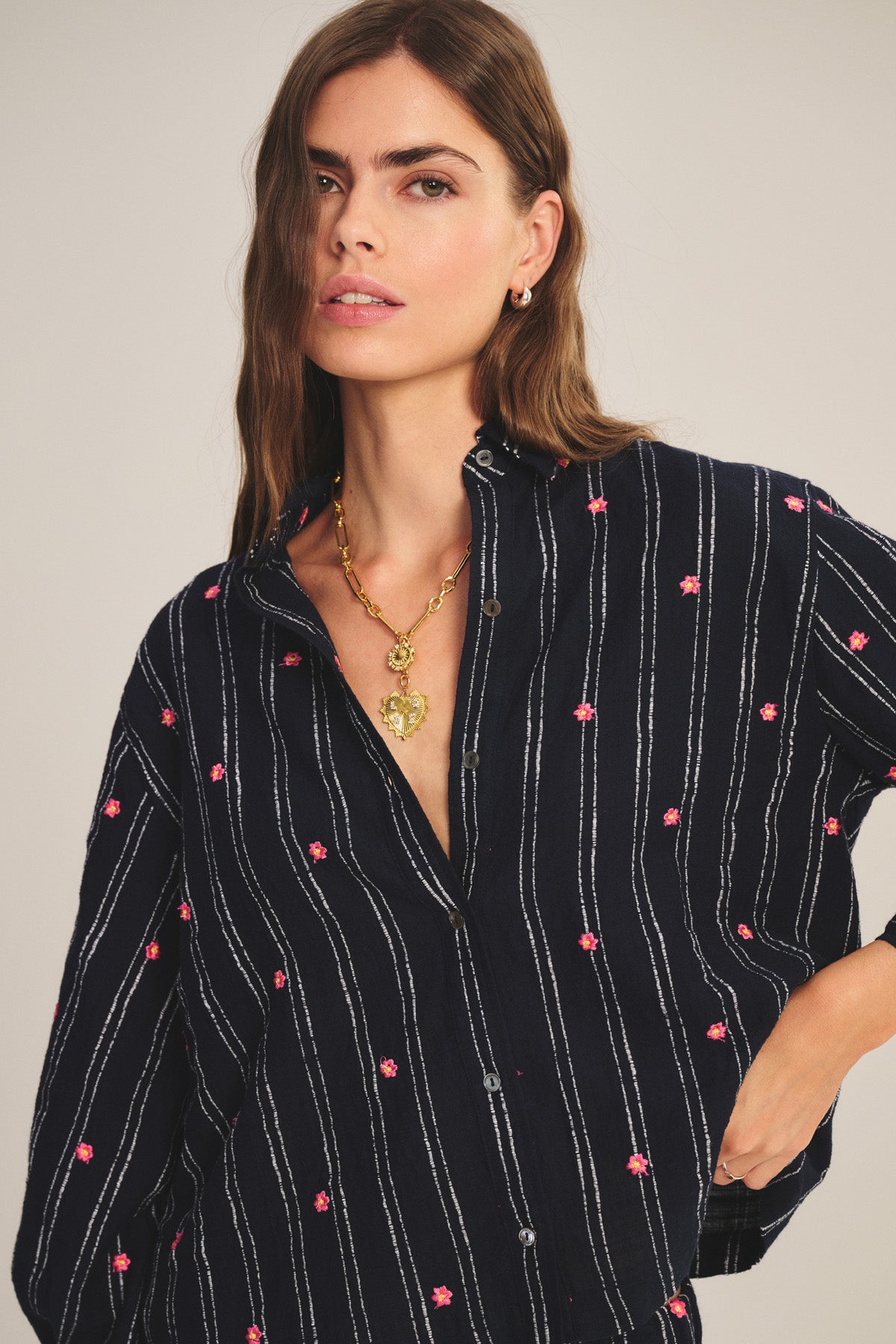 A woman with long brown hair wears the ASTREN SHIRT by Velvet by Graham & Spencer—a navy blue, pinstriped shirt with pink dots and embroidery—paired with a gold pendant necklace, standing against a plain background.