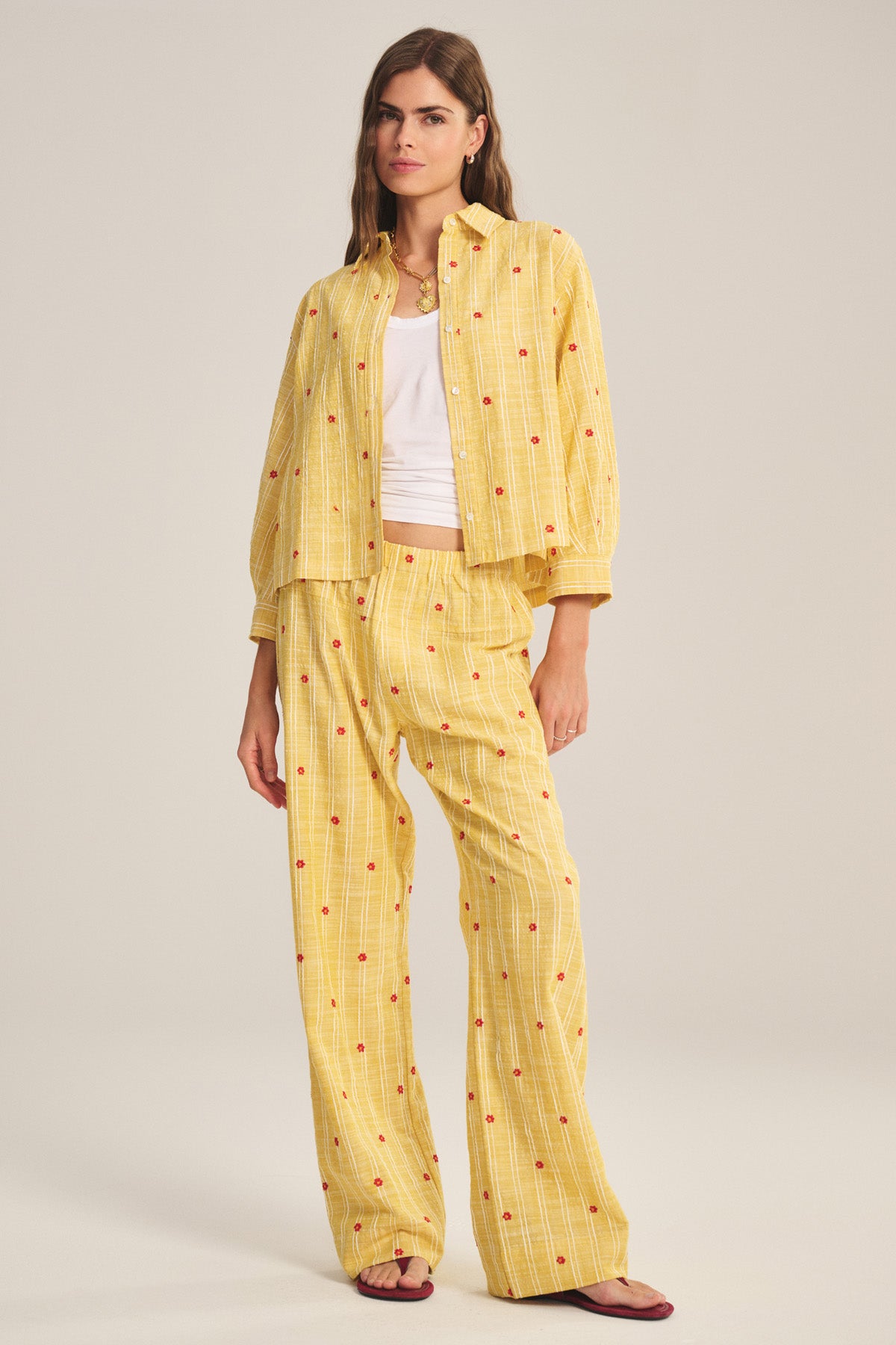 A woman wears the ASTREN SHIRT by Velvet by Graham & Spencer, a yellow embroidered top with matching wide-leg pants featuring small red dots, layered over a white tank top and paired with sandals.-41128024572097