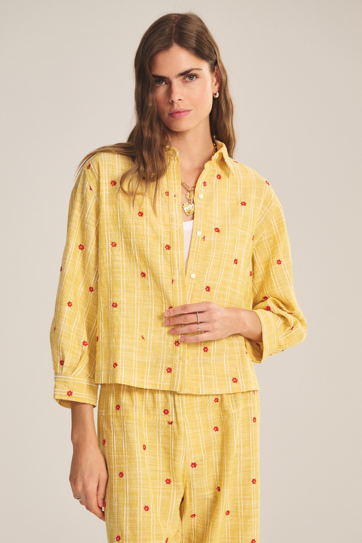 A woman wears the ASTREN SHIRT by Velvet by Graham & Spencer, a yellow checked embroidered shirt with small red flower patterns, paired with matching pants, standing against a plain background.-41128024637633